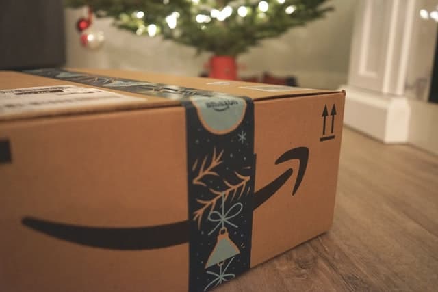 Small e-commerce delivery box with Amazon's arrow logo under a decorated Christmas tree.